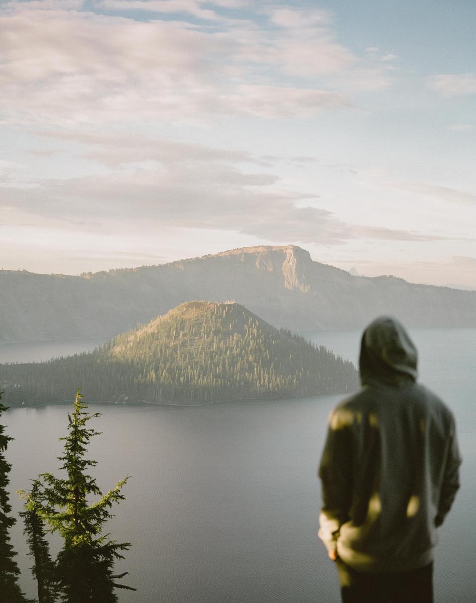 A person in a hoodie stands overlooking Crater Lake with a forested island and mountains in the distance.