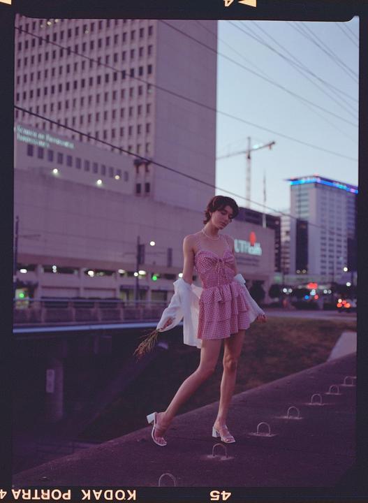 A woman in a pink gingham dress and white shirt stands on a ledge with city buildings in the background.