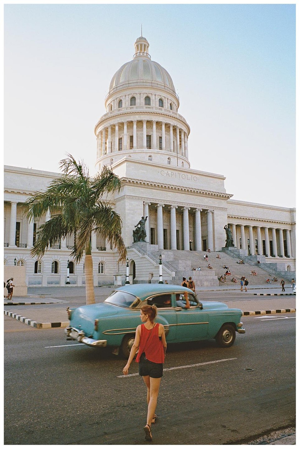 A woman walks in the street with a vintage car passing by and El Capitolio in the background.