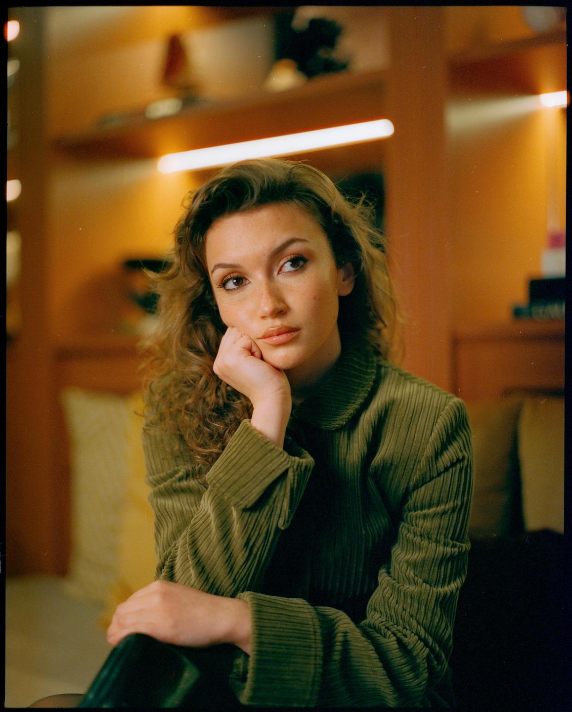 A woman with curly hair and a green corduroy jacket rests her chin on her hand while looking away from the camera.