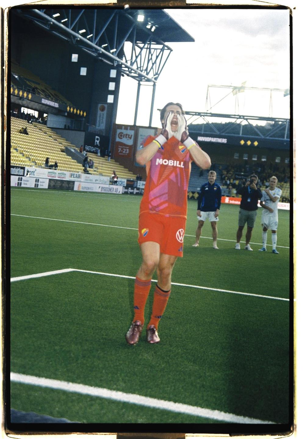 A soccer player in a red uniform shouts with his hands cupped around his mouth on a green field.