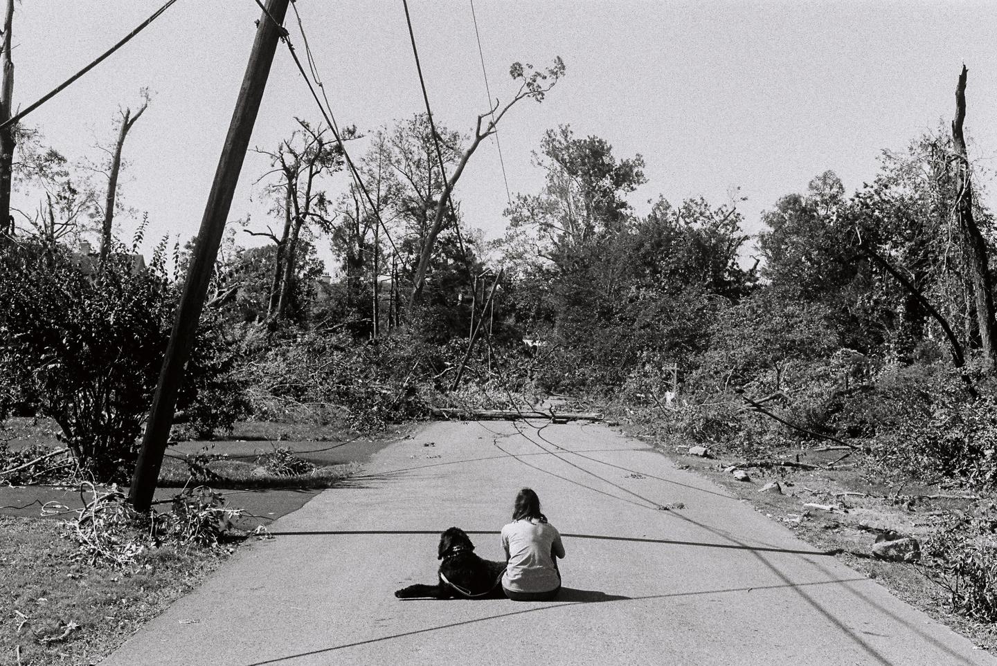 A woman and a dog sit on a road covered in debris from a tornado.