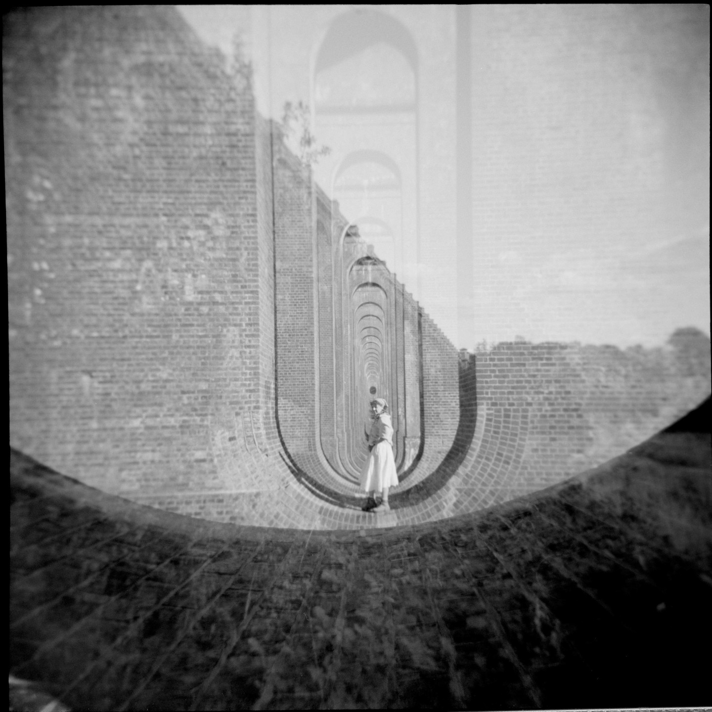A woman stands in a long brick archway with a double exposure of another archway above her.