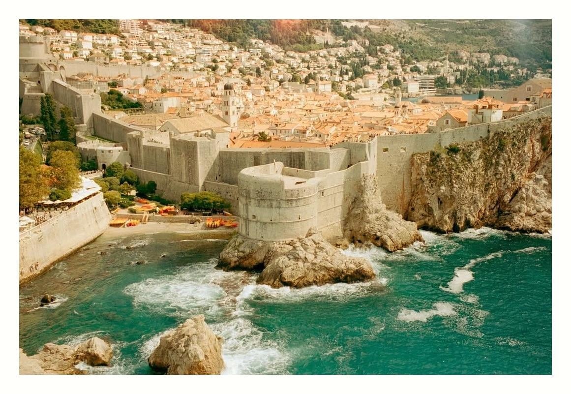 A fortified coastal city with red-tiled roofs is nestled against a hillside, with turquoise water crashing against its walls.