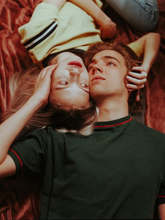 A couple lies on a red blanket, looking in opposite directions.