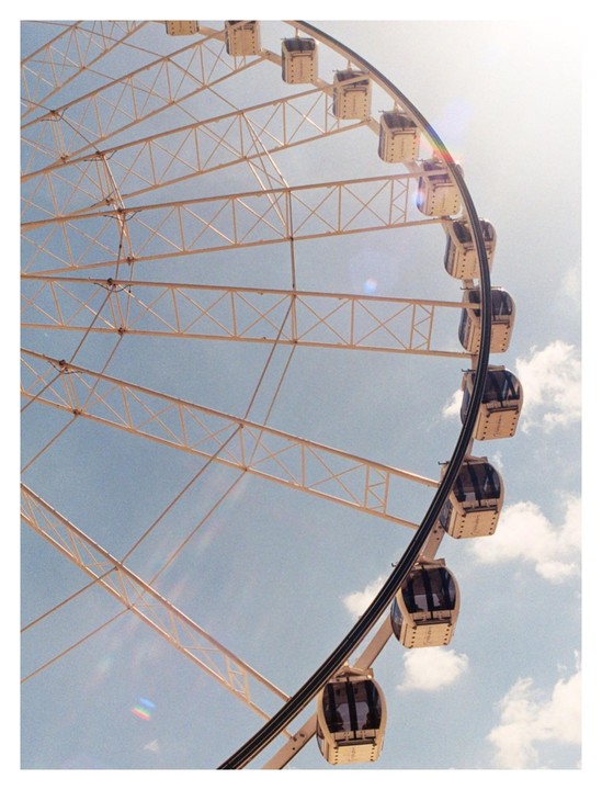 A large Ferris wheel with many gondolas is set against a bright blue sky with scattered clouds.