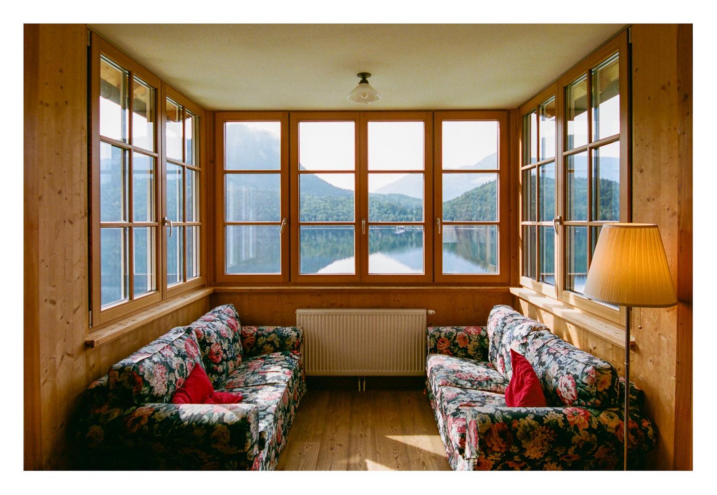 Two floral sofas with red pillows face each other in a wood-paneled room with large windows overlooking a lake and mountains.