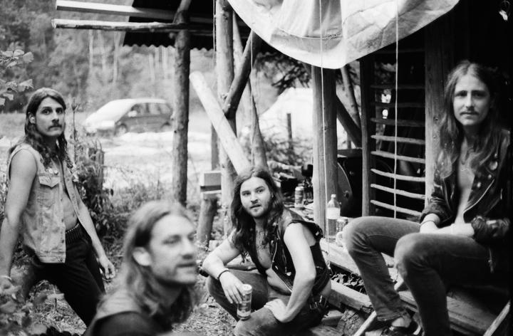 Four men with long hair are gathered outdoors, with one man in the foreground out of focus.