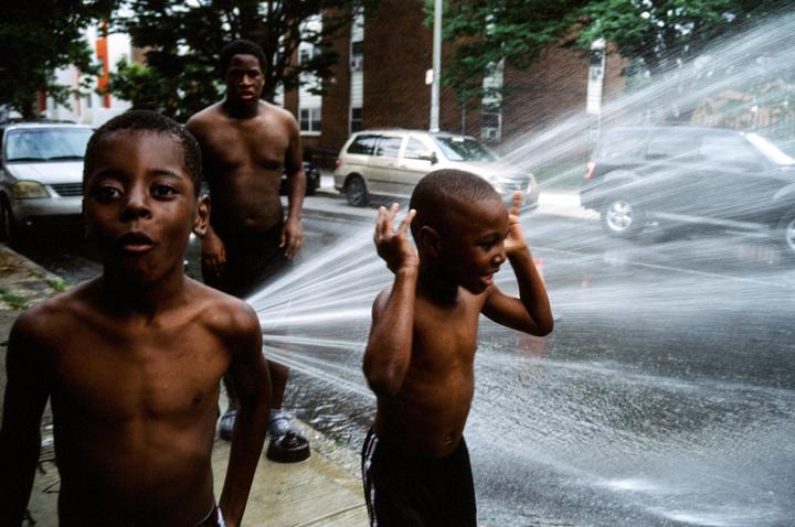 Three boys are playing in water spraying from a fire hydrant on a city street.