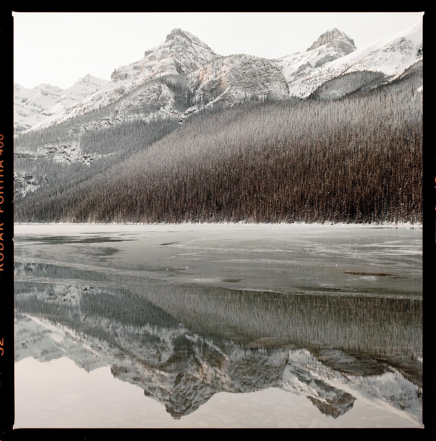 Snow-covered mountains and a forest reflect in a partially frozen lake.