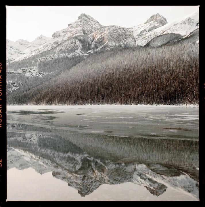 Snow-covered mountains and a forest reflect in a partially frozen lake.