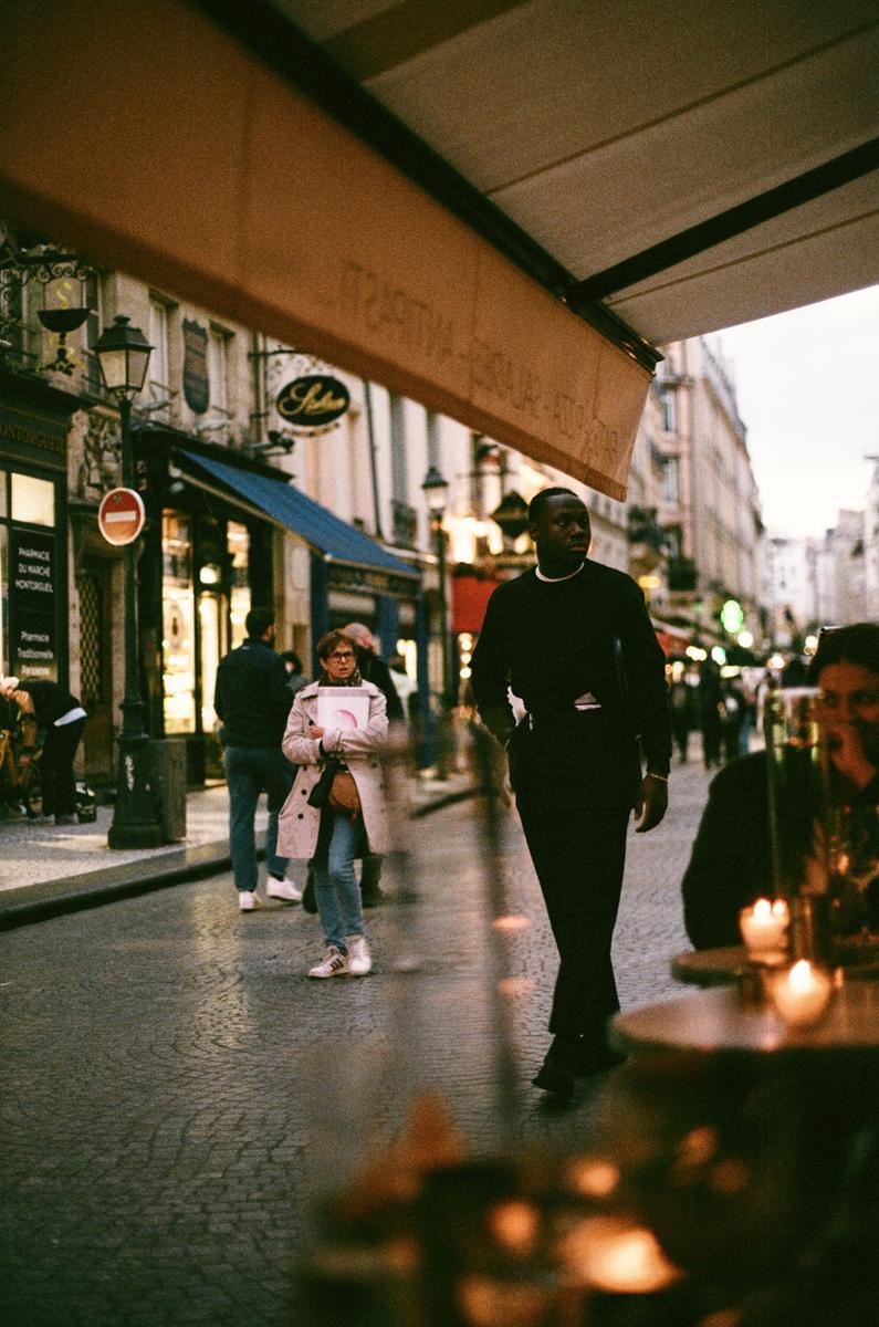A man walks down a cobblestone street past a cafe with an awning and a woman carrying a book.