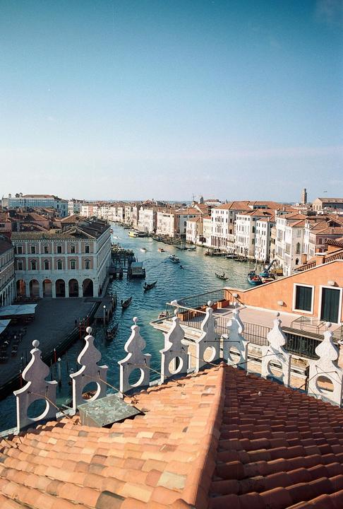 A high angle captures a canal with boats and buildings under a clear sky, with a tiled roof and decorative railing in the foreground.
