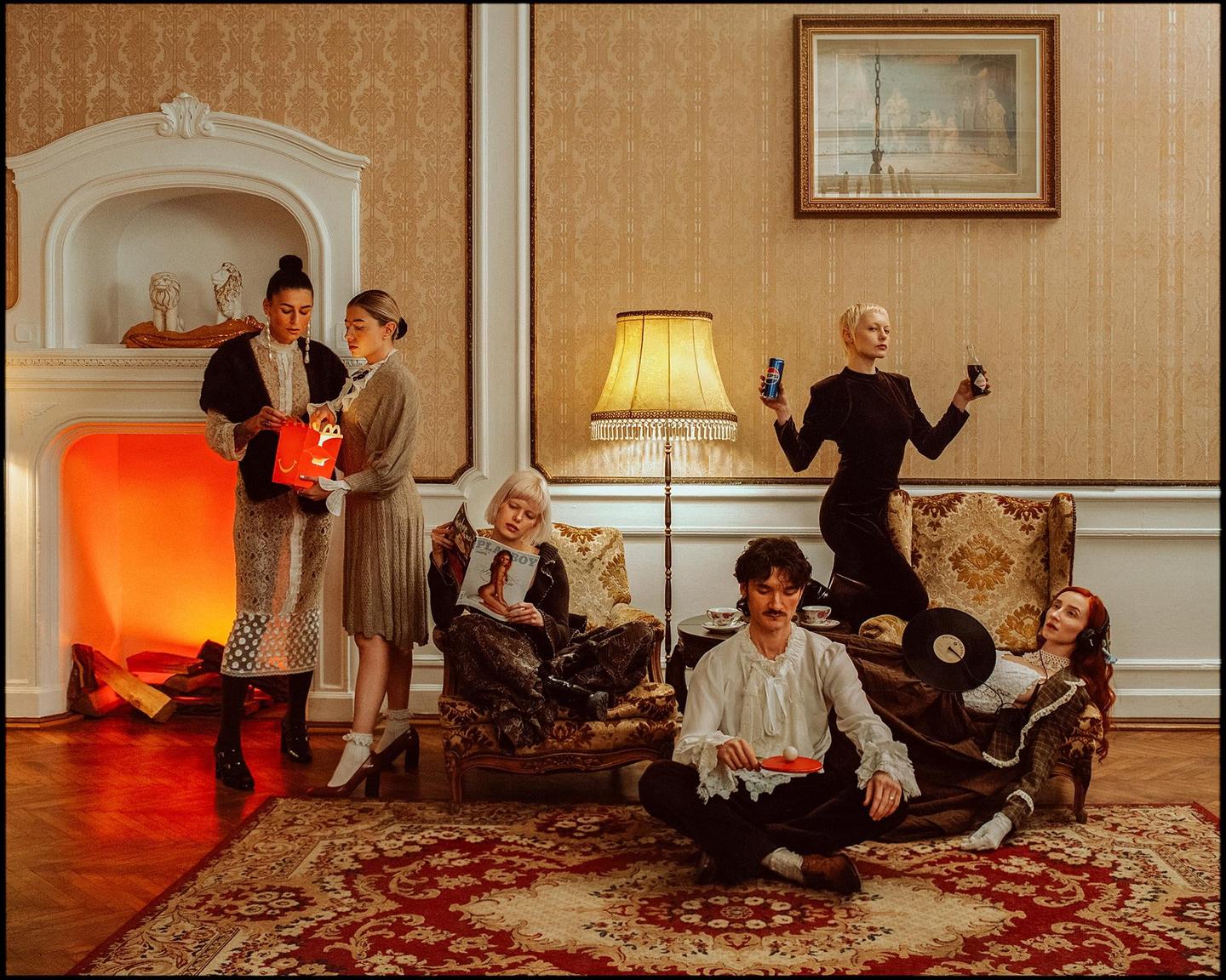 Six people are gathered in a room with ornate wallpaper and a fireplace, some holding drinks or magazines.