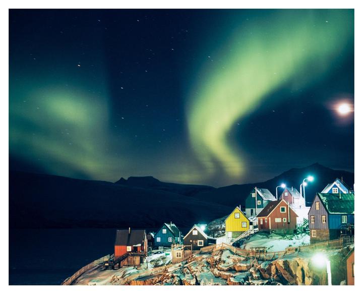 A vibrant aurora borealis illuminates a village of colorful houses nestled on a snowy hillside beside a body of water at night.