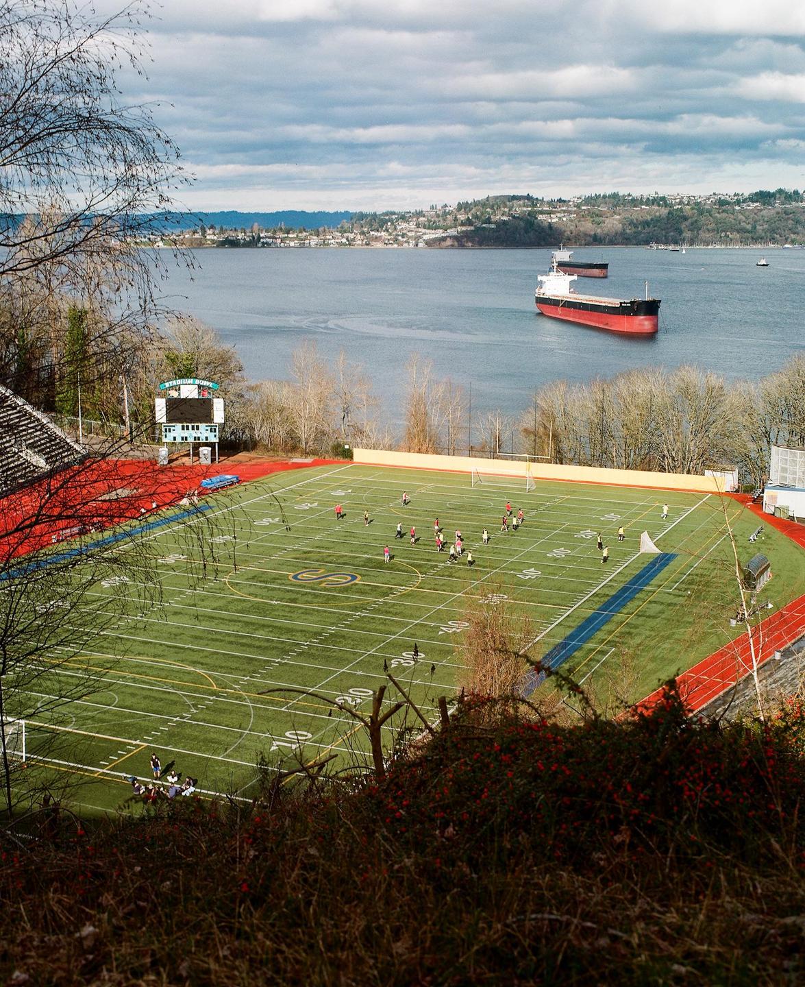 A football field with people playing is visible next to a body of water with ships and a distant town under a cloudy sky.