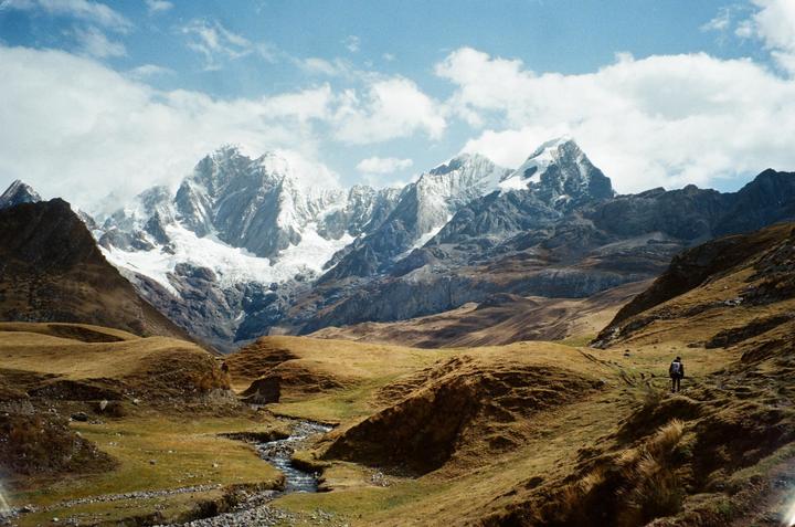 A person walks along a path in a valley with a stream, surrounded by snow-capped mountains under a cloudy sky.