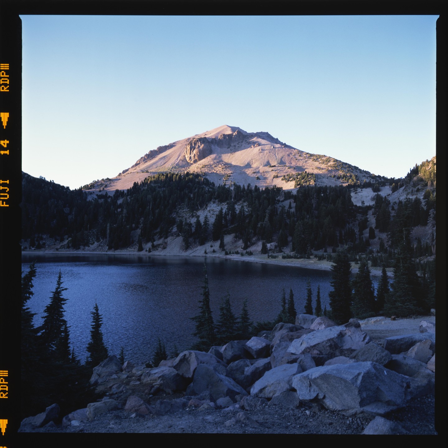 A mountain rises above a lake surrounded by pine trees and rocks.
