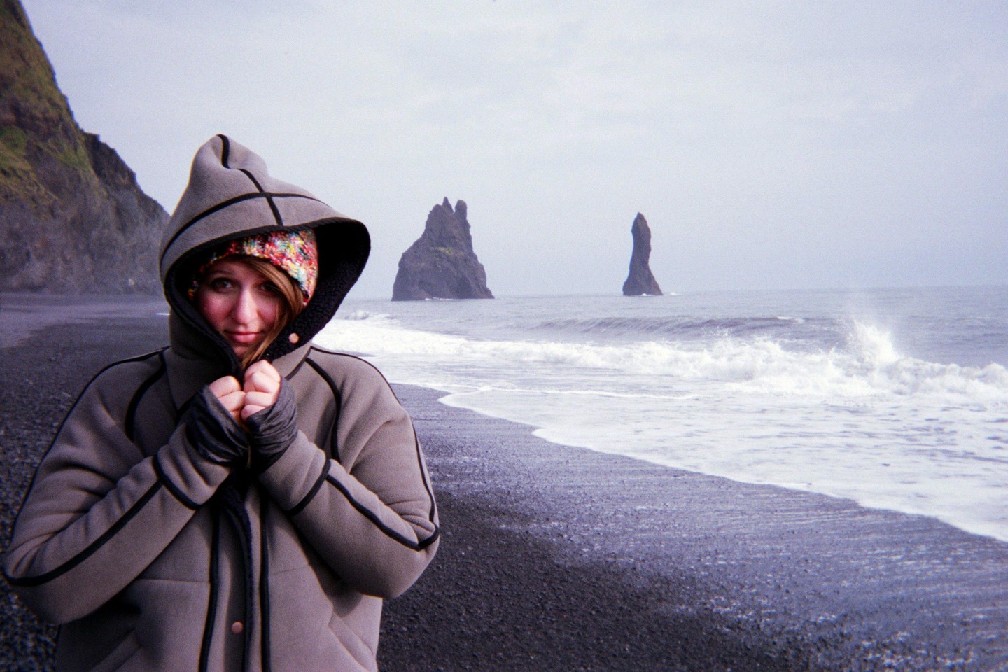 A woman in a hooded coat and colorful beanie stands on a black sand beach with rock formations in the ocean.