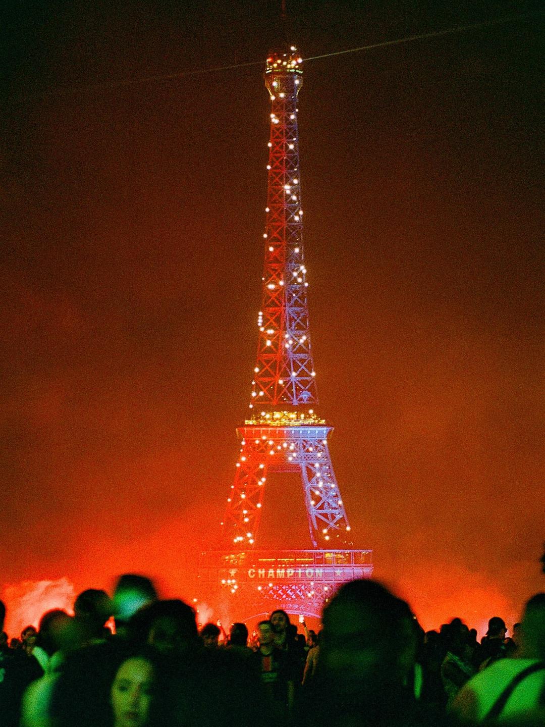 The Eiffel Tower is illuminated in red and blue lights with a crowd of people in the foreground.