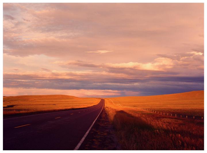 A long road stretches through a field under a dramatic sky at sunset.
