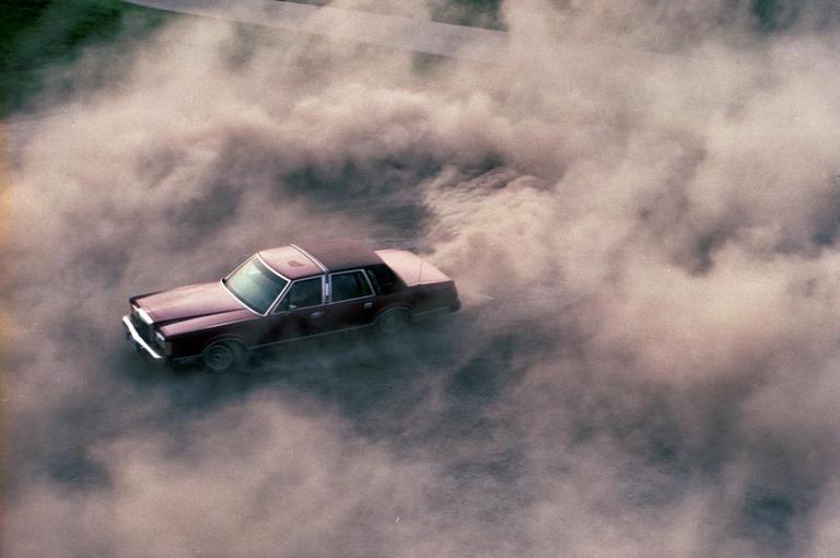 A maroon car is surrounded by a large cloud of dust.
