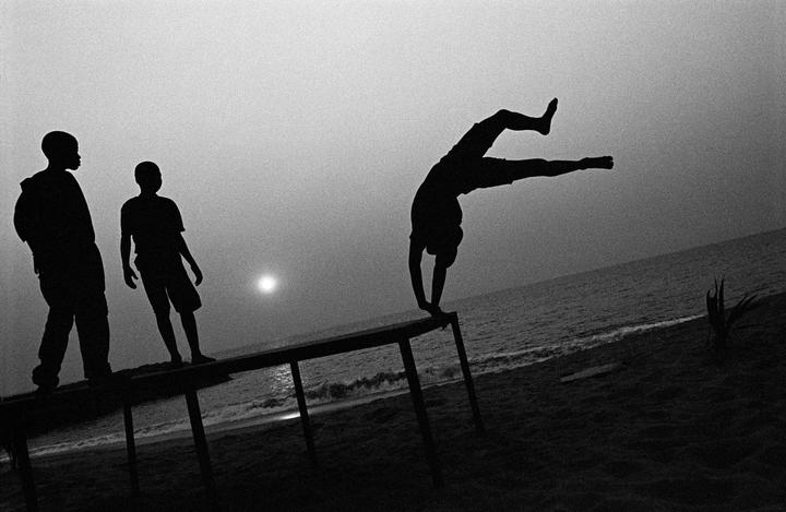 Three boys are silhouetted against the ocean and a bright sun, with one boy doing a handstand on a table.