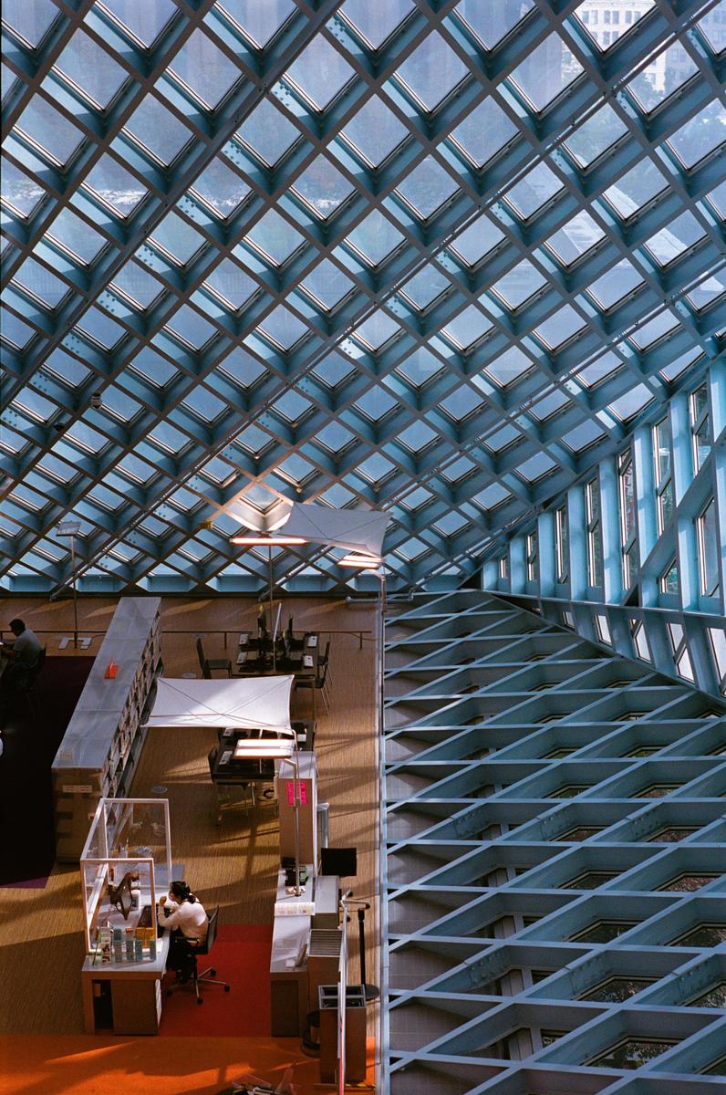 A woman works at a desk under a large glass and metal grid ceiling in a modern library.