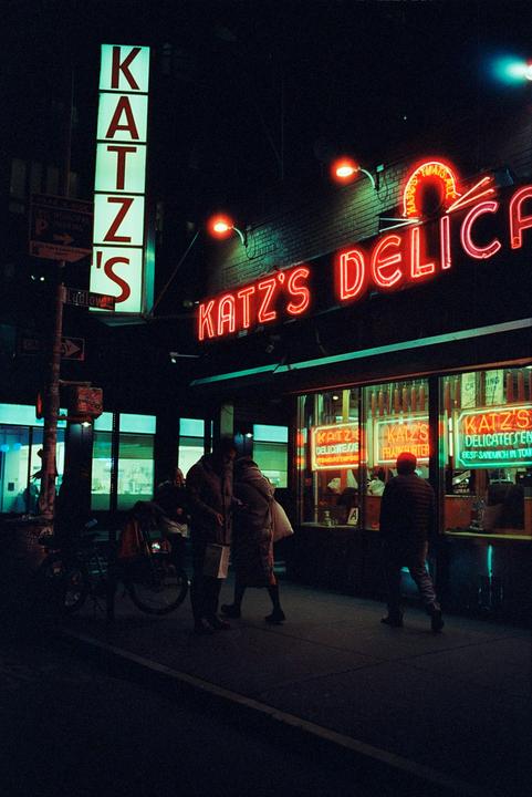 A neon sign for Katz's Delicatessen illuminates a street at night where people are walking.
