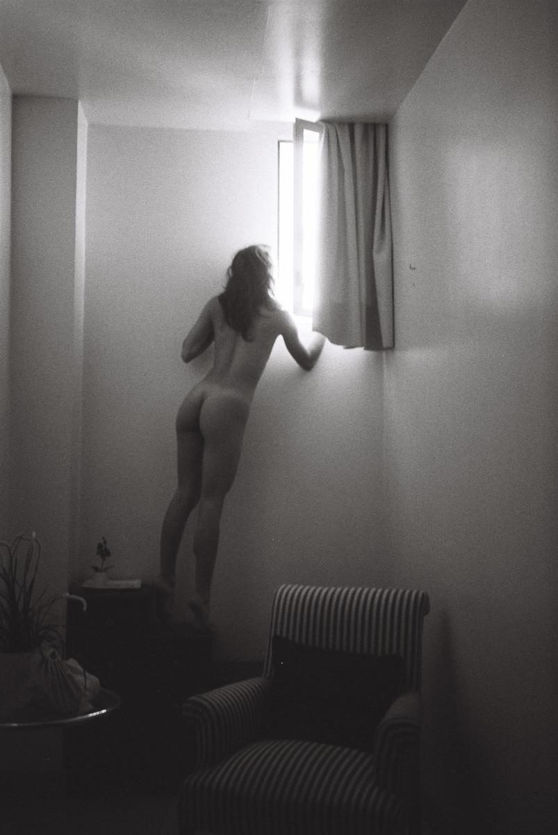 A nude woman stands on a piece of furniture to look out a window in a room with a striped armchair.