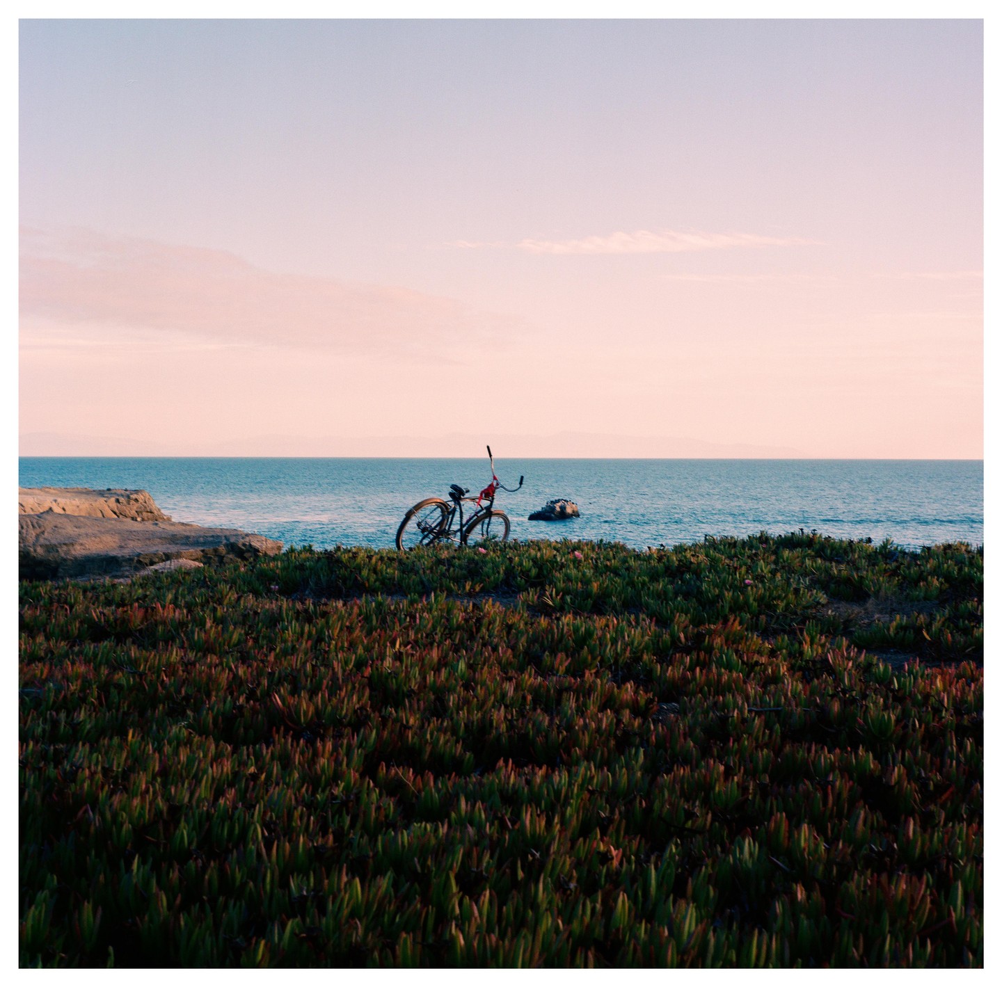 A bicycle is parked on a grassy cliff overlooking the ocean at sunset.