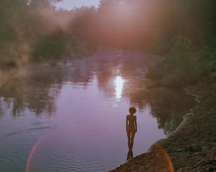 A woman stands in a river at dawn, with mist rising from the water and sunlight reflecting on the surface.
