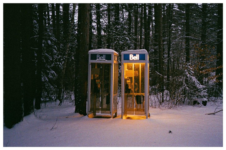 Two telephone booths stand in a snowy forest, one of them illuminated with a warm glow.