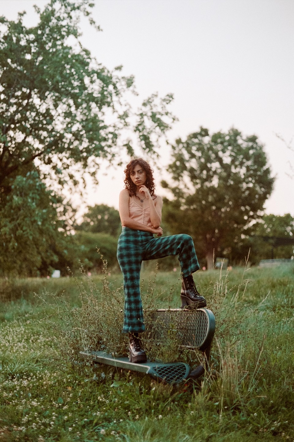 A woman in plaid pants and platform boots poses with one foot on a metal bench in a grassy field.