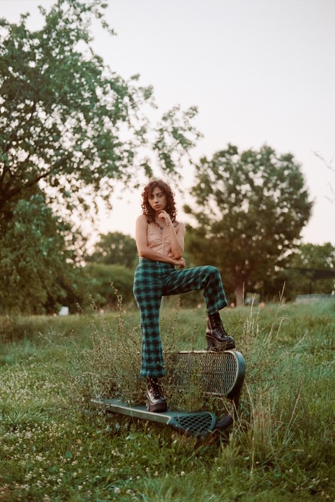 A woman in plaid pants and platform boots poses with one foot on a metal bench in a grassy field.