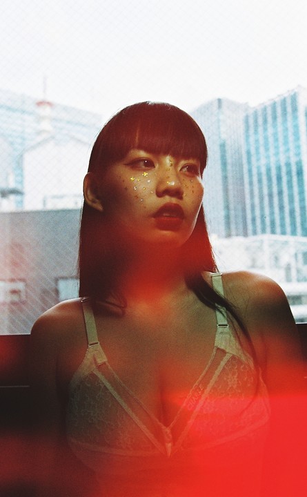 A woman with glitter on her face wears a bra and looks out a window at a city.