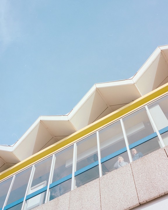 Two people are visible through the windows of a building with a zig-zag roofline.