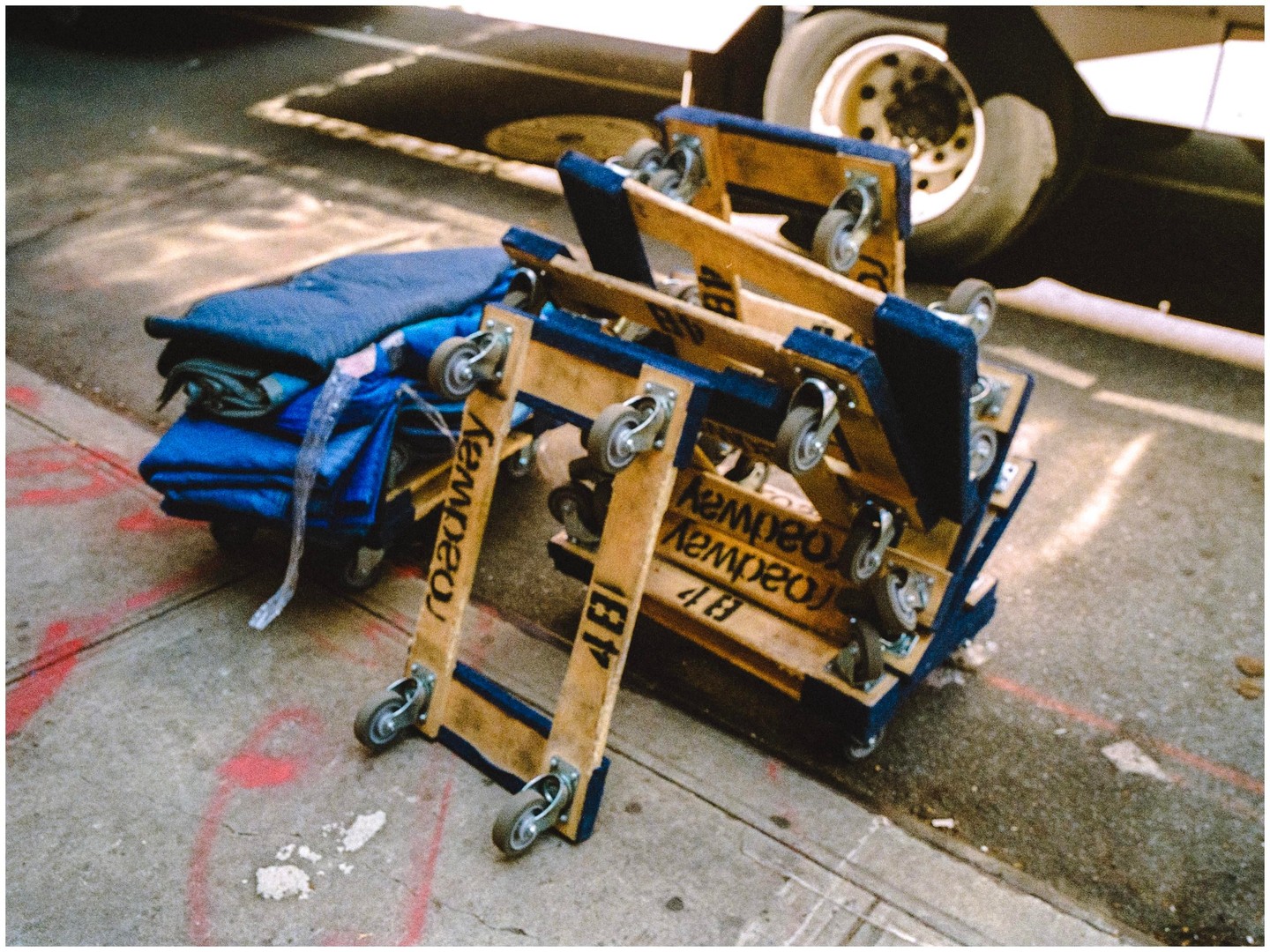 A stack of wooden moving dollies with blue padding and a pile of blue blankets are on a sidewalk next to a vehicle tire.