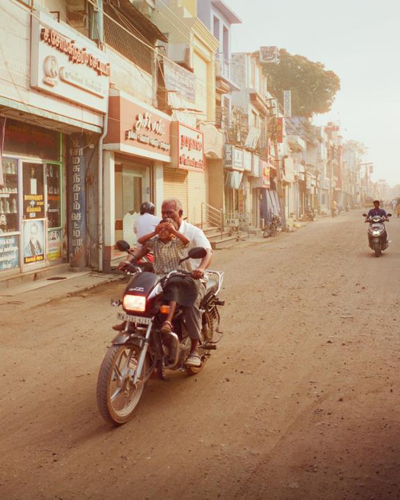 A man and a boy ride a motorcycle down a dusty street lined with buildings.