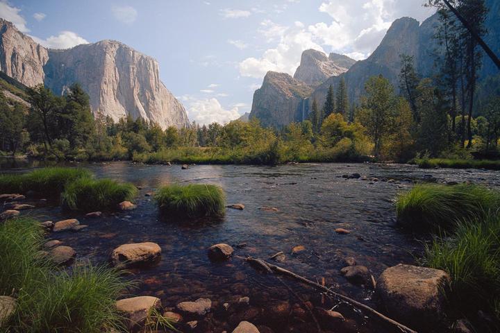 A river with rocks and grass in the foreground flows towards a valley with mountains and trees under a cloudy sky.