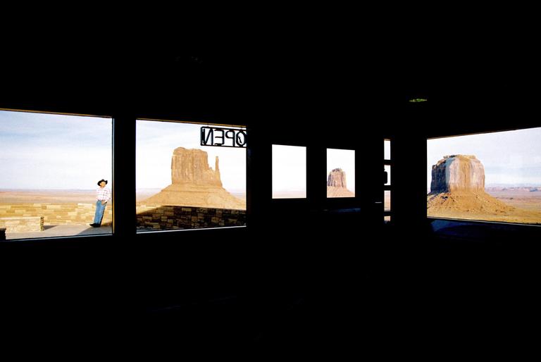 A man in a cowboy hat stands outside a window looking at a desert landscape with large rock formations.