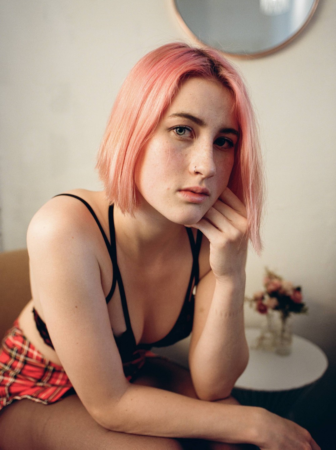 A woman with pink hair and a red plaid skirt poses for a portrait indoors.
