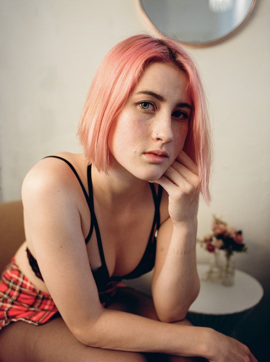A woman with pink hair and a red plaid skirt poses for a portrait indoors.