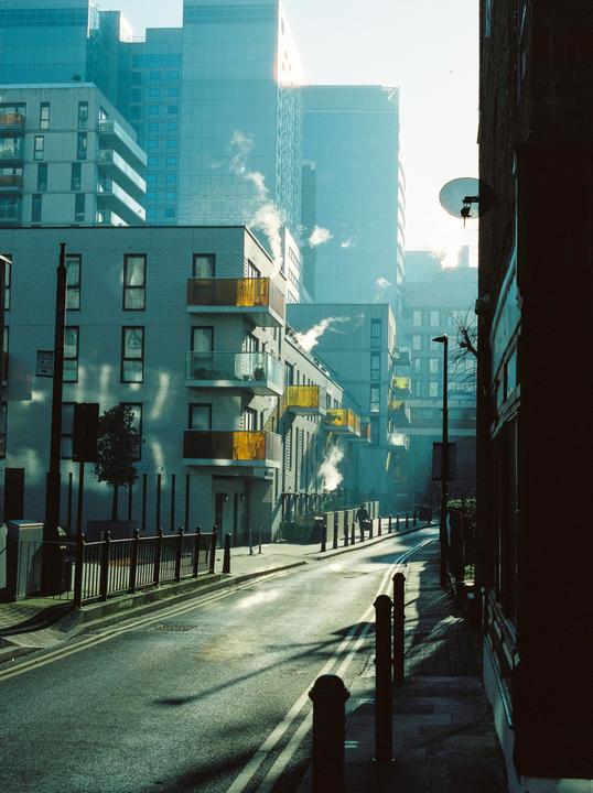 A street in East London is lined with modern buildings, some with yellow balconies, and steam rises from several places.