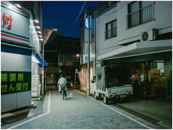 A person rides a bicycle down a narrow street at night, with buildings and a truck visible on either side.