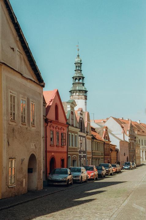 A row of colorful buildings with parked cars lines a cobblestone street, leading to a tall church tower under a clear sky.