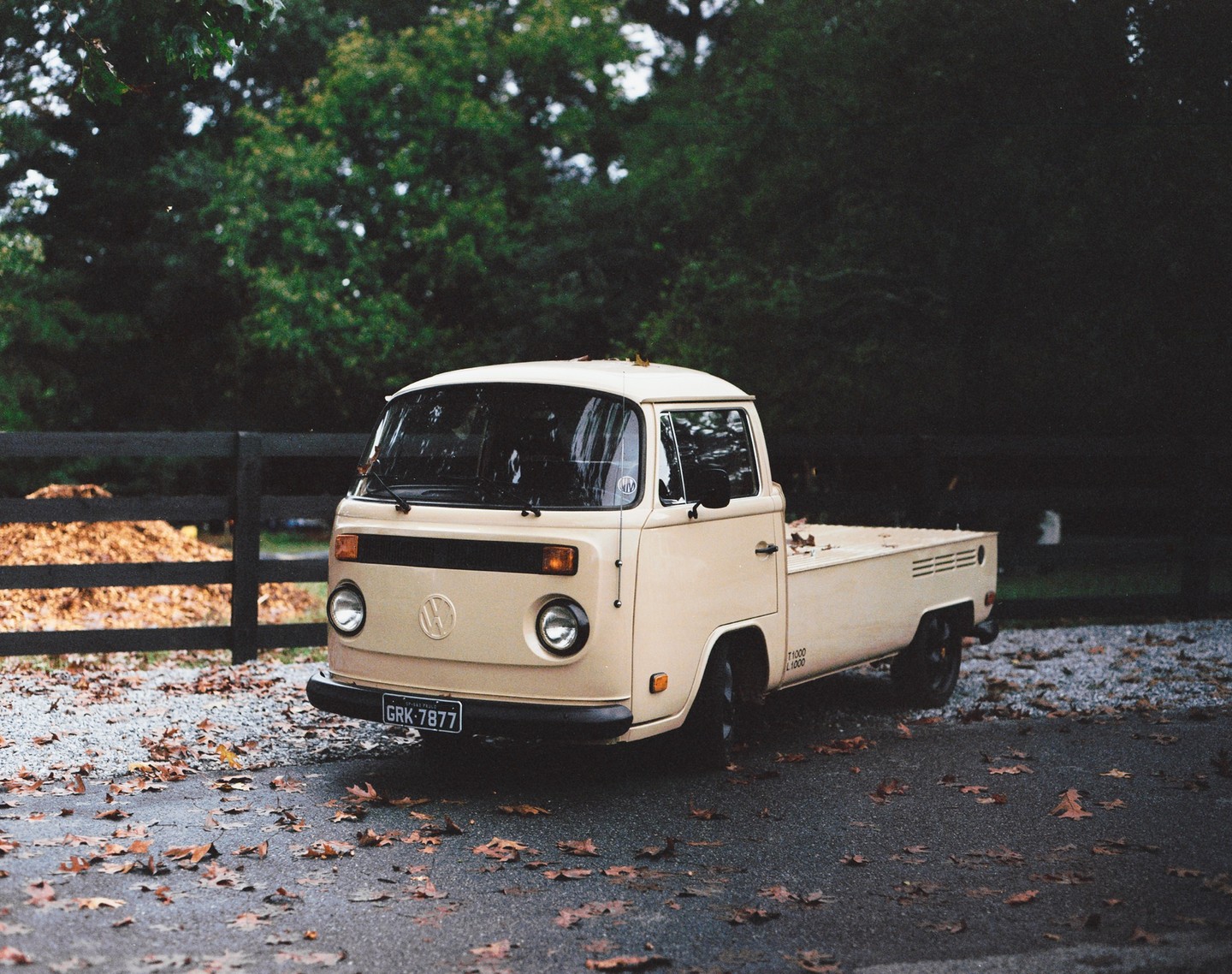 A beige Volkswagen pickup truck is parked on an asphalt surface covered with autumn leaves, with a wooden fence and trees in the background.