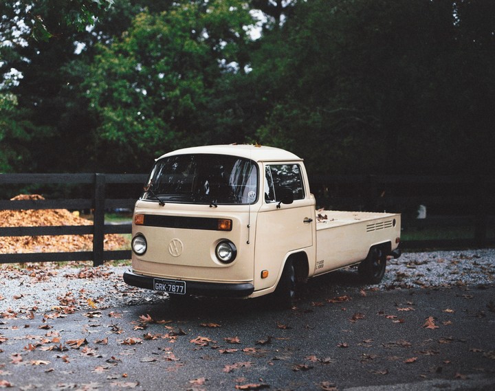 A beige Volkswagen pickup truck is parked on an asphalt surface covered with autumn leaves, with a wooden fence and trees in the background.