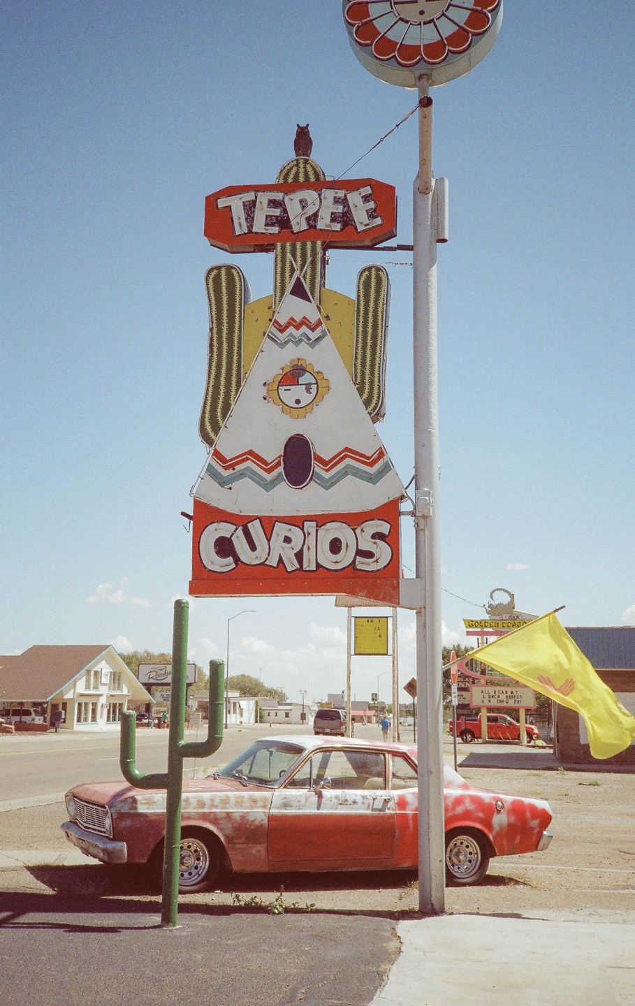 A red and white car is parked next to a large sign for "Tepee Curios" with a cactus sculpture in the foreground.