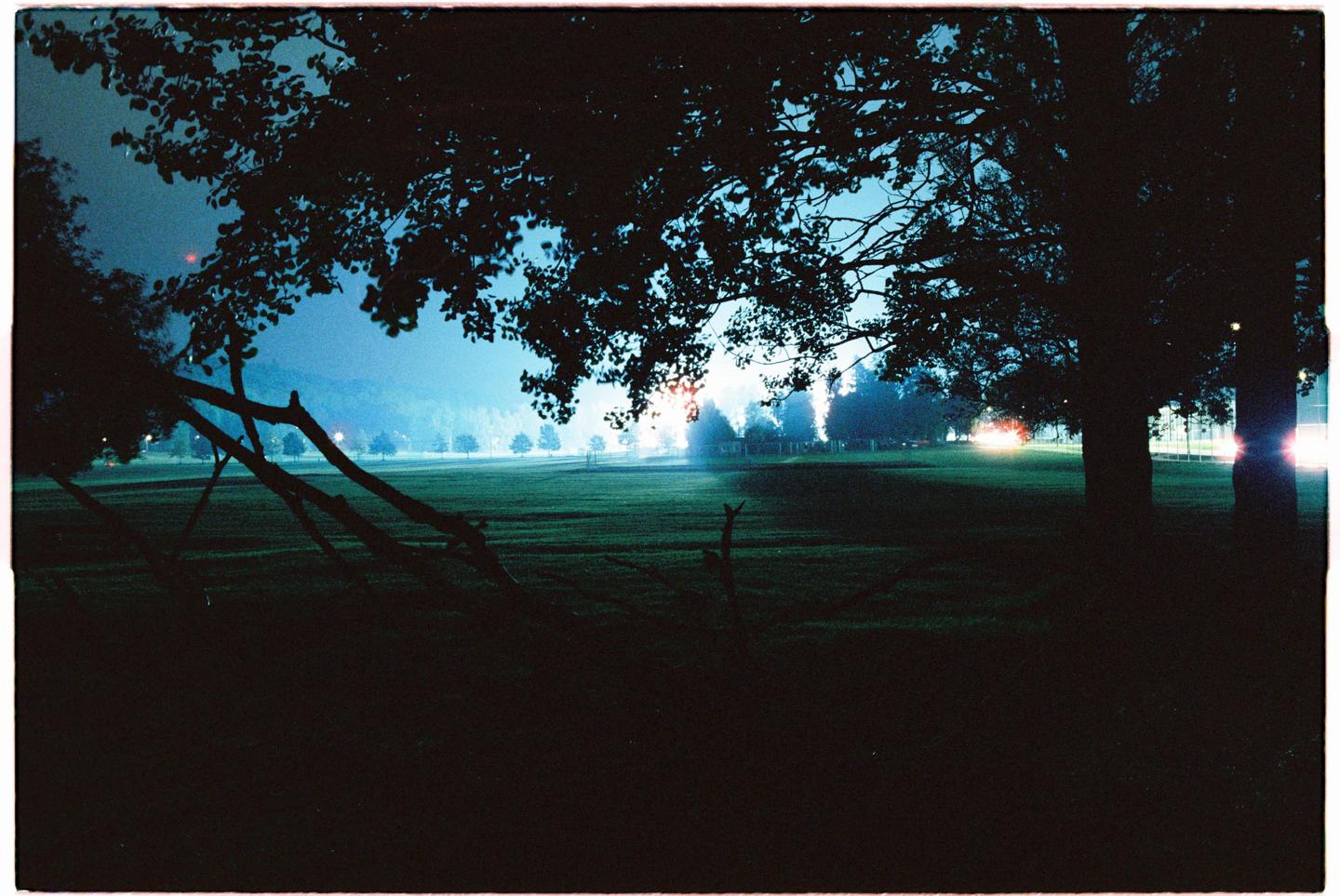A dark field is illuminated by distant lights under a blue sky with silhouetted trees in the foreground.